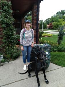 Emily stands by her house and wears a black and white striped shirt, jeans, and a baseball cap. She is holding the leash of a black Great Dane. She has three suitcases next to her.